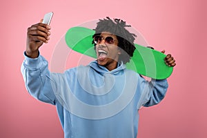 Excited black teenager with skateboard deck taking selfie on smartphone over pink studio background