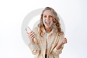 Excited blond girl winning online money prize, shouting yes with joy and triumph, celebrate victory with mobile phone in