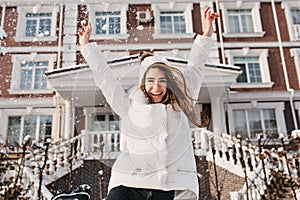 Excited brightful emotions of joyful pretty young woman expressing soround snowfall on street in winter time on house