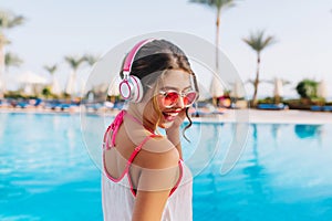 Excited brunette girl with tanned skin listening favoring music while sunbathing by the open-air pool. Portrait of