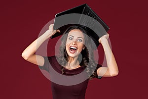 Excited business woman in maroon dress holding notebook or computer over head on red background.