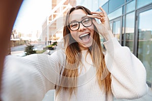 Excited business woman posing walking outdoors near business center wearing eyeglasses take selfie by camera