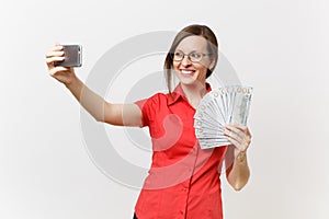 Excited business woman in red shirt doing taking selfie shot on mobile phone with bundle lots of dollars, cash money