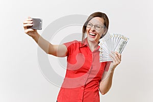 Excited business woman in red shirt doing taking selfie shot on mobile phone with bundle lots of dollars, cash money