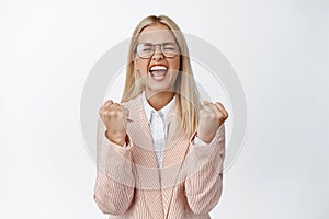 Excited businesswoman shouting and clenching fists, triumphing and celebrating success, standing in suit and glasses