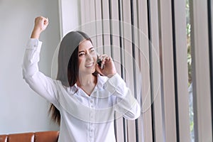 Excited businesswoman talking with smartphone holding in her hands while raised hands up celebrate with good news in office