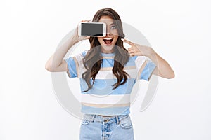 Excited carefree smiling woman hold smartphone horizontal cover one eye with mobile phone, pointing cellphone display