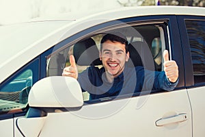 Excited casual guy sitting behind the steering wheel of the car showing thumbs up gesture as has passed the exam and obtain driver photo