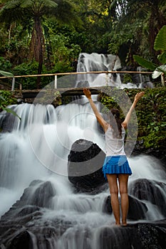 Excited Caucasian woman raising arms in front of waterfall. View from back. Jembong waterfall. Singaraja, Bali, Indonesia