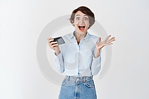 Excited caucasian woman screaming happy and surprised, holding mobile phone and triumphing, winning online or in video