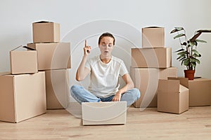 Excited Caucasian woman wearing white t shirt sitting on floor surrounded with cardboard boxes with belongings and working on