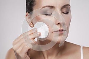 Excited cheerful confident lovely beautiful mature woman is using cotton pad with micellar water for removing make up from face,