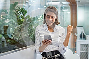 Excited cheerful young business woman with fist up receiving good news on smart phone and screaming in office