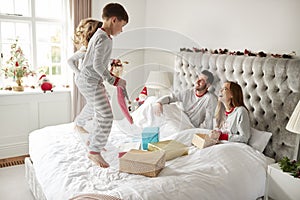 Excited Children Jumping On Parents Bed At Home As Family Open Gifts On Christmas Day