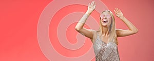Excited chilling energized young blond woman in silver stylish glittering dress sunglasses raise hands up having fun