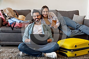 Excited couple using laptop to book tickets and hotel online, preparing for vacations and packing suitcases