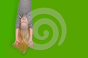 Excited crazy little blonde girl hanging happy upside down hands up over isolated green studio background. Emotion