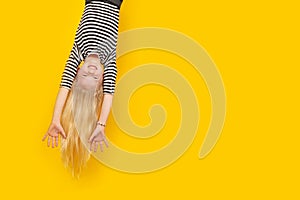 Excited crazy little blonde girl hanging happy upside down hands up over isolated yellow studio background. Emotion