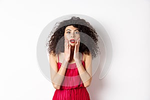 Excited curly-haired woman touch face with disbelief, looking amazed at camera, staring at promo, standing in red ress