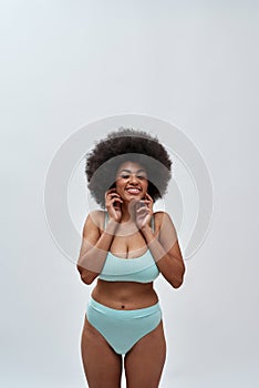 Excited curvy young woman with afro hair style wearing blue underwear smiling at camera, posing  over light