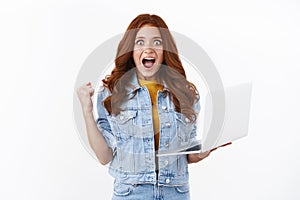 Excited cute redhead girl in denim, hold laptop, pump her fist joyfully, rejoicing excellent news, stare camera winning