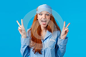 Excited cute redhead girl in sleep mask and pyjama showing peace signs and screaming thrilled overwhelmed positive
