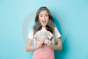 Excited cute woman winning money, holding dollar bills and smiling amazed, got fast credit, standing over blue