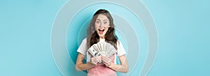 Excited cute woman winning money, holding dollar bills and smiling amazed, got fast credit, standing over blue