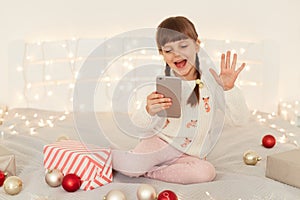 Excited dark haired female kid with pigtails holding mobile phone in hands having video call or livestream, waving hand to smart