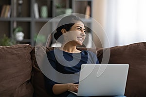 Excited dreamy Indian woman distracted from laptop, sitting on couch