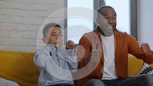 Excited emotional african american dad and son watching football game on tv, enjoying goal, tracking shot, slow motion