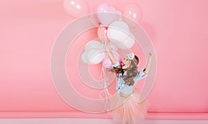 Excited entertainment of happy little girl in tulle skirt running with flying balloons isolated on pink background