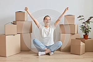 Excited extremely happy woman wearing white T-shirt sitting on the floor near cardboard boxes with stuff, celebrating relocation