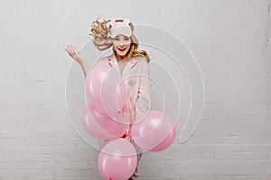 Excited fair-haired girl in pink pajamas holding bunch of helium balloons. Indoor portrait of inspired young lady in