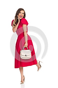 Excited Fashionable Woman In Red Dress, Gold High Heels And Beige Purse Is Standing On One Leg