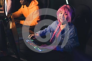 Excited female gamer rookie in pink wig spending her free time in PC bang playing on desktop computer using headphones