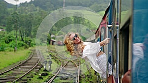 Excited female tourist leans out train door in rich green tropics. Curly-haired woman in sunglasses enjoys scenic rail