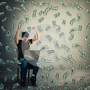 Excited freelancer guy seated working on laptop raising hands up holding fists as celebrating win. Money falling like dollar rain