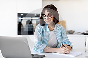 Excited freelancer lady working with laptop in kitchen interior and taking notes, writing down information