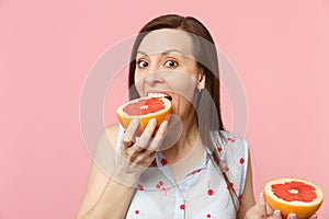 Excited funny young woman in summer clothes holding in hand, biting half of fresh ripe grapefruit isolated on pink