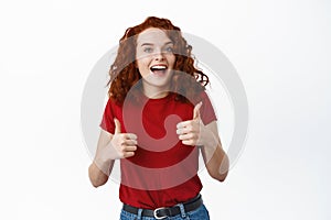 Excited ginger girl praising good work, showing thumbs up and smiling satisfied, nod in approval and say yes, give