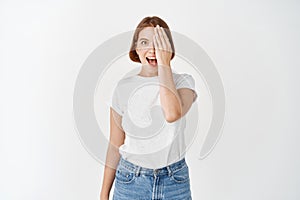 Excited girl cover half of face and eye, gasping amazed and looking at something awesome, standing in t-shirt and jeans