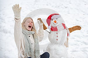 Excited girl plaing with a snowman on a snowy winter walk. Making snowman and winter fun. Cute snowman in hat and scalf