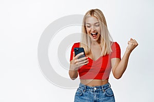 Excited girl winning on smartphone, holding phone and scream with joy, triumphing, making money on cellphone, white