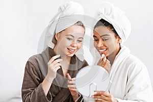 Excited girlfriends in bathrobes putting lipsticks together, looking at mirror