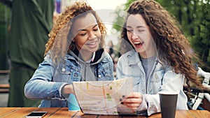 Excited girls tourists are checking city map and looking around then laughing sitting in street cafe in foreign country