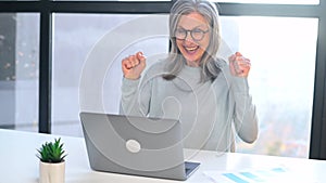 Excited gray-haired senior woman looking at laptop screen sitting at desk in office