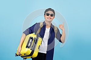Excited handsome Asian male tourist in casual clothes hat backpack holding suitcase bag, shows thumb up gesture on blue background