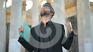 Excited handsome Middle Eastern man with digital tablet rejoicing success standing on sunny urban city street talking