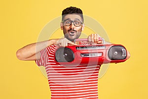 Excited happy bearded man in striped t-shirt and fashionable eyeglasses holding in hand red tape recorder, pushing play button